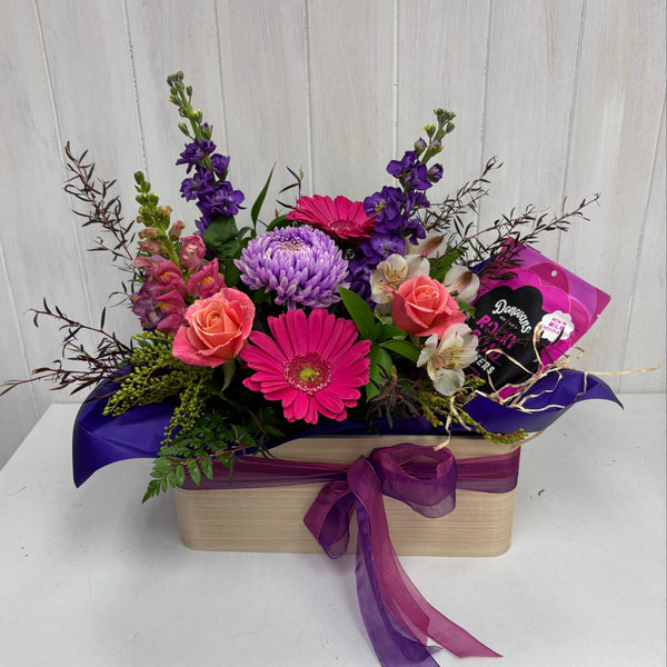 Colorful flower arrangement in a box with purple ribbon on a white surface.